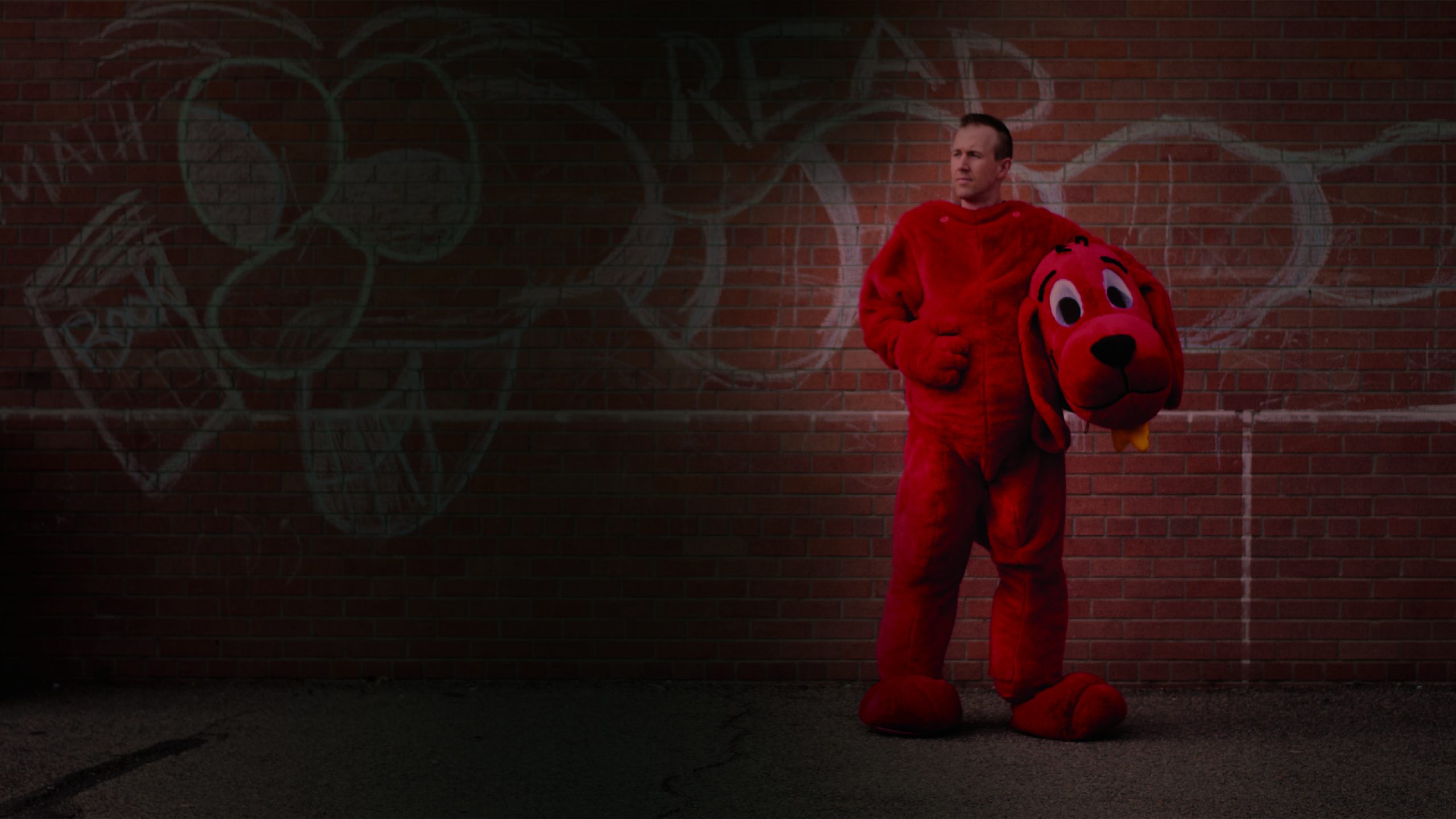 Ron S. Doyle standing against a graffitied brick wall in a Clifford the Big Red Dog costume, holding the costume head under one arm.